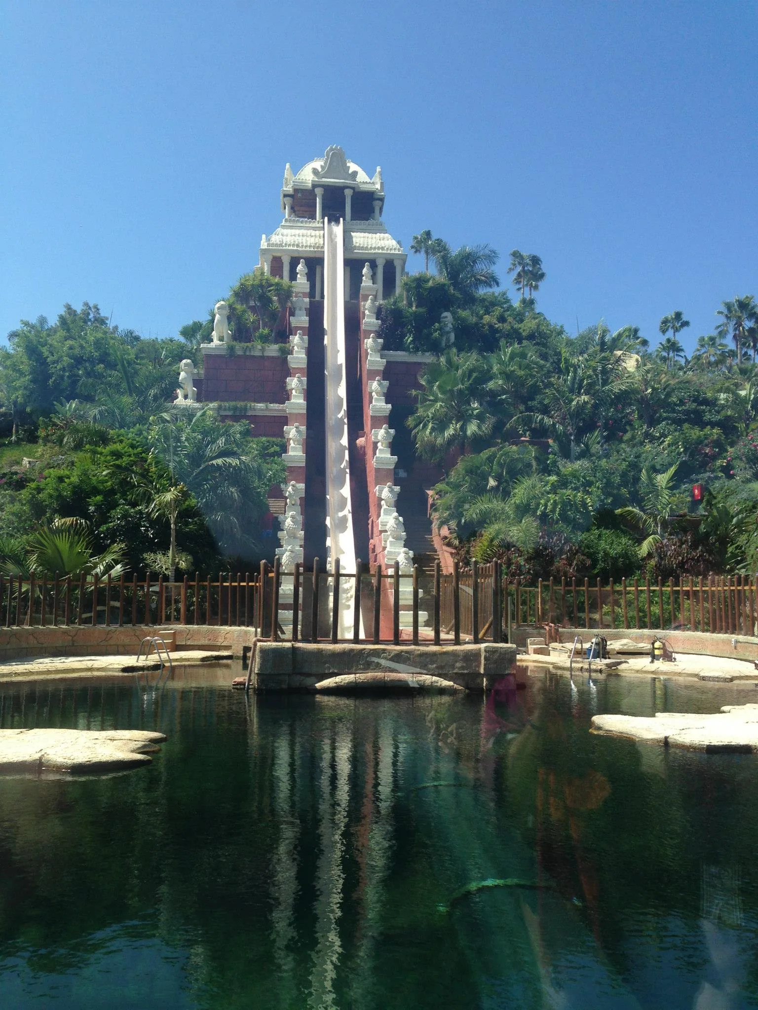 A tall water slide reflected in a pool beneath a blue sky